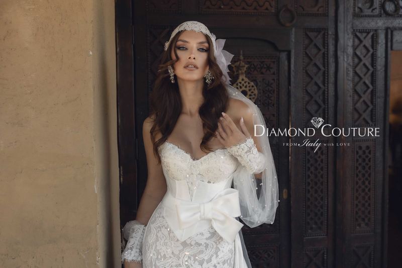 Abito da sposa Diamond Couture 2027: corpetto a cuore in pizzo, grande fiocco in raso e velo con perle, davanti a una porta intagliata