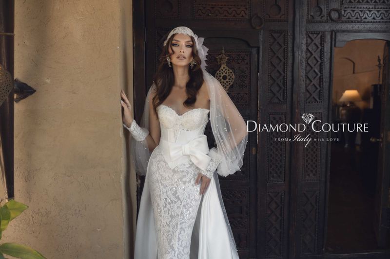 Abito da sposa Diamond Couture 2027: in pizzo senza spalline, fiocco in vita e sovragonna in raso, davanti a una porta intagliata