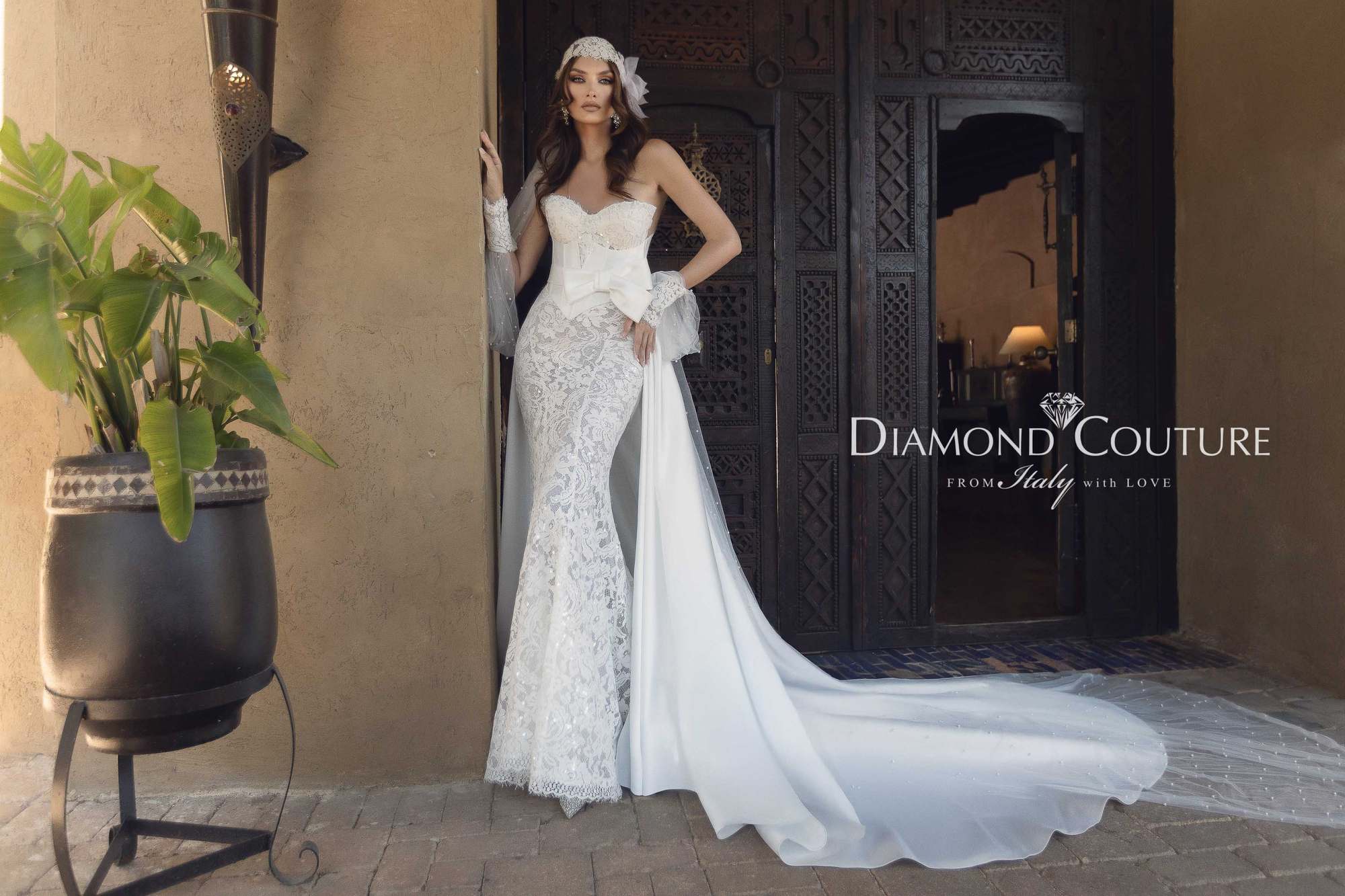 Abito da sposa Diamond Couture 2027: a figura intera, sirena in pizzo con fiocco e strascico in raso, davanti a una porta intagliata