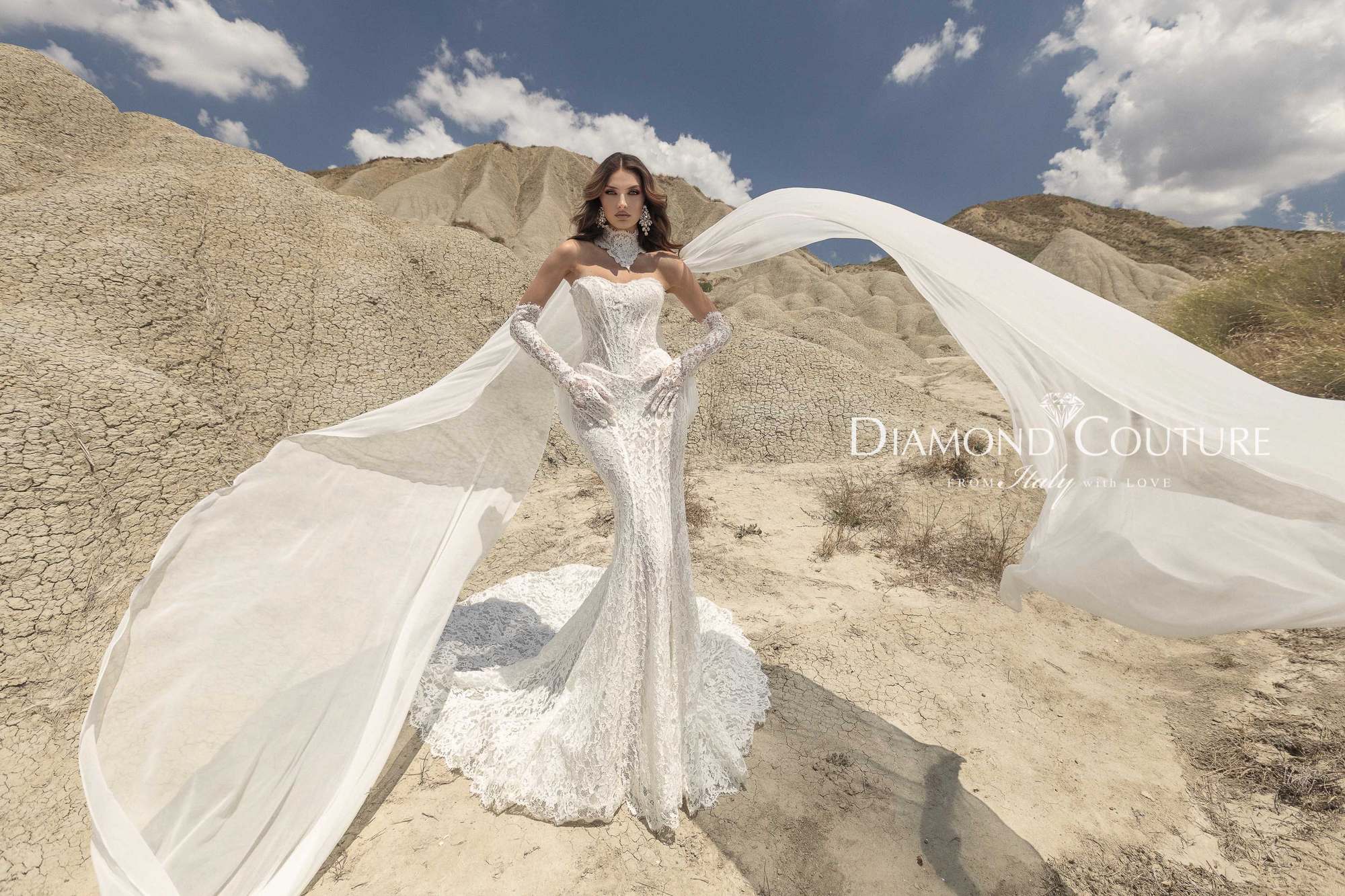 Abito da sposa Diamond Couture 2027: a figura intera, sirena in pizzo a fascia con guanti lunghi e teli leggeri al vento, tra i calanchi