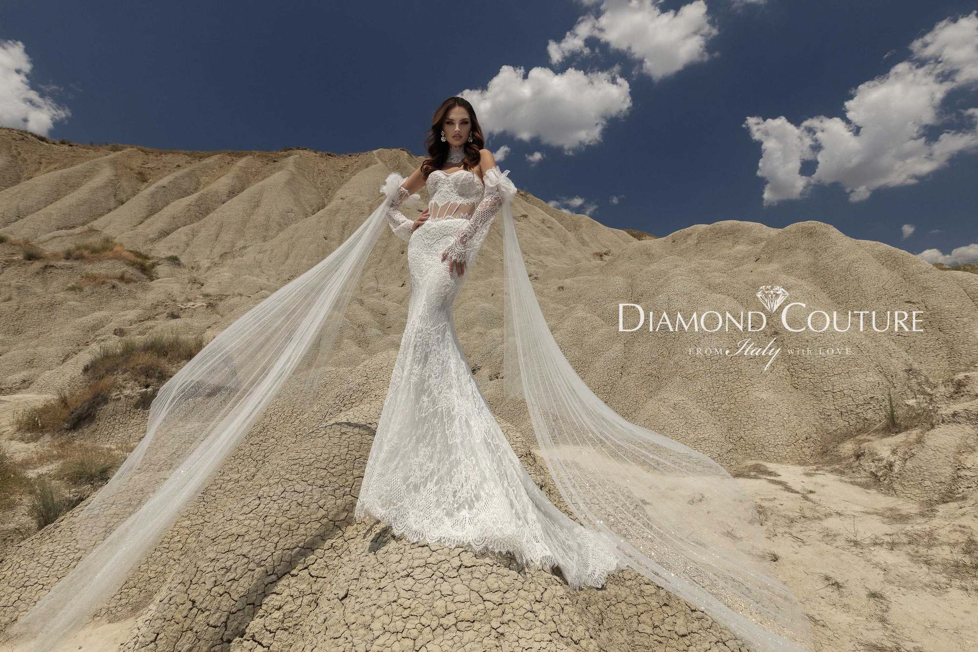Abito da sposa Diamond Couture 2027: a figura intera, sirena in pizzo con orlo smerlato e lunghi veli dalle maniche, sui calanchi