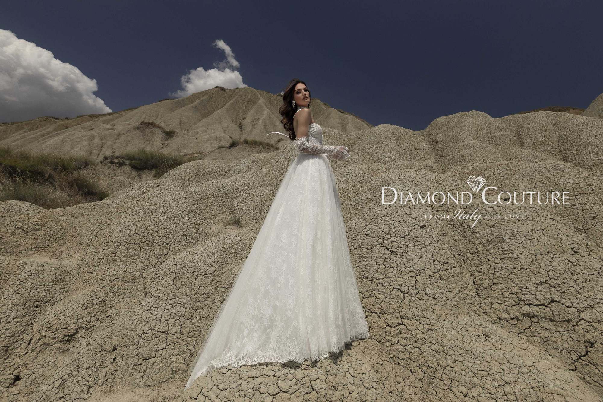 Abito da sposa Diamond Couture 2027: vista laterale, gonna ampia in pizzo con strascico e guanti lunghi, tra i calanchi sotto il cielo